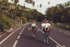 two people riding bikes down the middle of a road with palm trees in the background