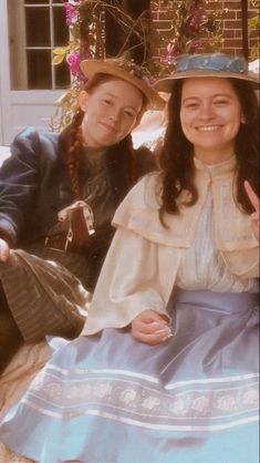 two women dressed in historical clothing sitting next to each other on a bench and smiling at the camera