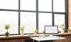 a desk with a computer on top of it next to a window filled with potted plants