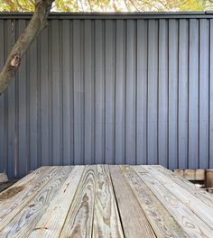 an empty wooden table sitting in front of a metal wall with a tree next to it