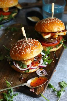 two hamburgers with onions, tomato and lettuce on a cutting board next to some dipping sauce