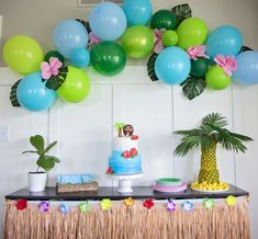 a table topped with a blue cake and lots of green, pink and blue balloons