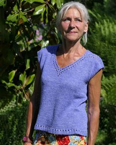 an older woman with white hair and blue shirt standing in front of some bushes, looking at the camera