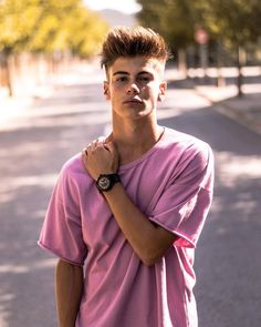 a young man in a pink shirt is standing on the street with his hand under his chin