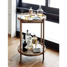 a bar cart with drinks on it sitting in front of a window next to a wooden floor