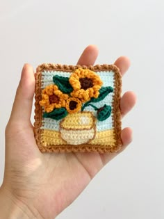 a hand holding a small crocheted square with flowers on it and a vase in the middle