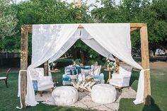 an outdoor seating area with white drapes and pillows on the grass, surrounded by trees