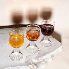 three wine glasses sitting on top of a stone slab with different colored liquids in them