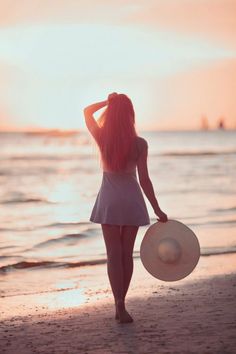 a woman with long pink hair is walking on the beach holding a large white hat