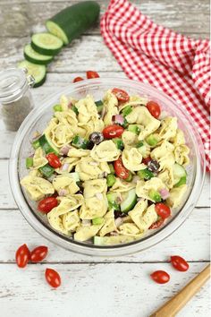 a glass bowl filled with pasta salad next to cucumbers and tomatoes on a white wooden table