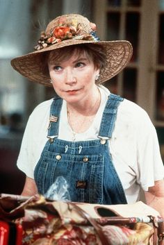 a woman wearing overalls and a hat standing in front of a pile of stuff