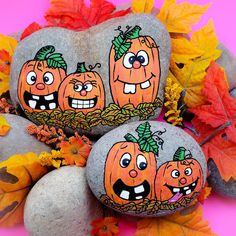 three pumpkins painted on rocks surrounded by leaves and autumn foliage, with faces drawn on them