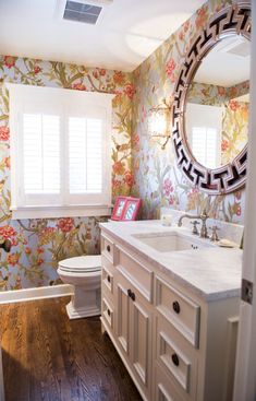 a bathroom with floral wallpaper and wooden floors is seen in this image, the mirror above the sink has an interesting design