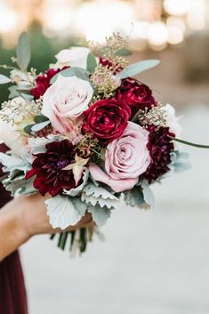 a woman holding a bouquet of red and pink flowers with greenery in her hand