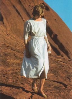 a woman in a white dress is walking up a hill with her hand on her hip