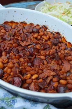 a white dish filled with beans and meat on top of a blue table cloth next to a bowl of coleslaw