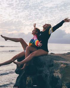 two young women sitting on top of a rock near the ocean with their arms outstretched