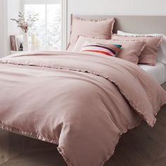 a bed with pink comforter and pillows in a white room next to a window