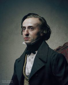 a man with long hair wearing a black suit and yellow tie sitting in a chair