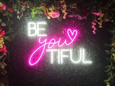 a neon sign that says be you tiful with flowers and greenery in the background