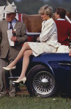 two women sitting on the back of an old fashioned car with men standing behind them