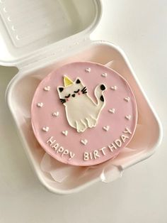 a pink birthday cake with a cat on it in a plastic container next to a white box