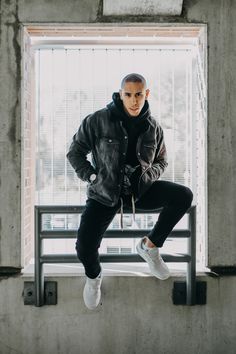 a man in black jacket and white sneakers sitting on a window sill with his hands on his hips