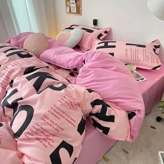 a bed covered in pink sheets and pillows with black letters on the comforter, next to a window