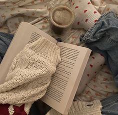 an open book and some sweaters are laying on the bed next to a cup of coffee