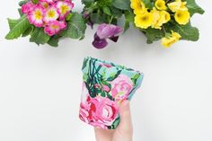 a person holding up a flower pot with pink and yellow flowers in it next to green plants