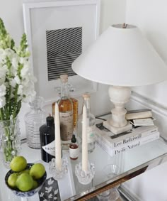 a glass table topped with books and bottles next to a lamp on top of a dresser