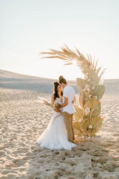 a bride and groom standing on the beach in front of a palm tree with their arms around each other