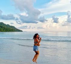 a woman standing on top of a sandy beach next to the ocean holding a cell phone