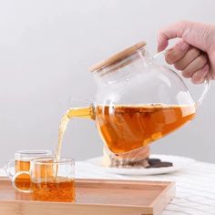 someone pouring tea into a glass pitcher on a tray with two cups next to it