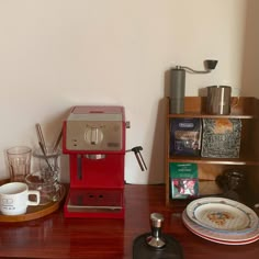 a red coffee maker sitting on top of a wooden table next to a cup and saucer