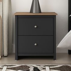 a black night stand with two drawers and a lamp on top, next to a bed