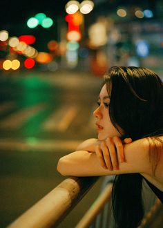 a woman leaning on a railing looking at the street lights in the distance with her hand on her shoulder