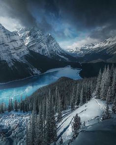 snow covered mountains and pine trees under a cloudy sky with a lake in the foreground