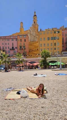 a woman laying on top of a sandy beach next to buildings and palm trees in the background