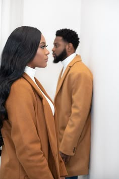 a man and woman standing next to each other in front of a white wall, looking at each other