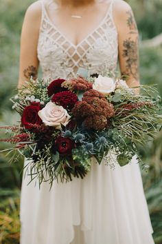 a woman holding a bouquet of flowers and greenery in her hands, wearing a wedding dress