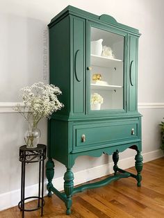 a green china cabinet sitting on top of a hard wood floor next to a vase with flowers