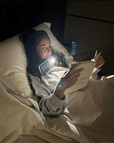 a woman laying in bed reading a book and holding an electronic device to her ear