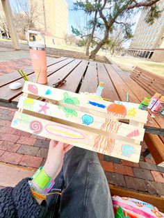 a person sitting on a bench holding up a piece of wood with different colored designs