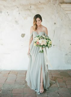 a woman standing in front of a white wall wearing a dress and holding a bouquet