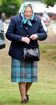 an older woman wearing a blue coat and plaid skirt carrying a black handbag while standing in the grass