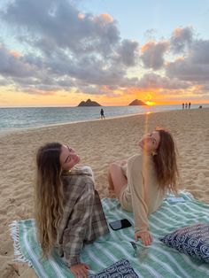 two women sitting on a towel at the beach watching the sun set over the ocean