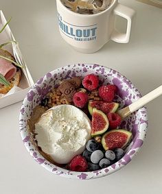 a bowl filled with fruit and ice cream on top of a white table next to a mug