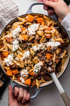 two hands holding a skillet full of pasta with meat, cheese and carrots