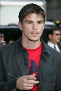 a young man in a red shirt and black jacket is looking at the camera with his hand on his hip